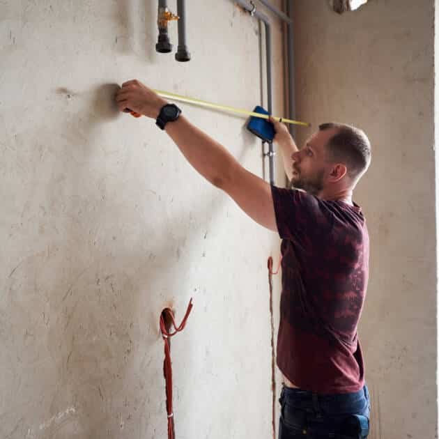 Side view of serious man using measuring tape in room with underfloor heating pipes. Designer in shirt taking measurements while working on renovation of apartment.