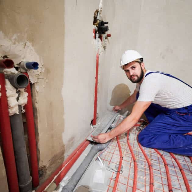 Portrait of bearded young man in safety helmet installing pipes in apartment. Male plumber in work overalls installing water trap in the shower. Concept of home renovation and plumbing works.