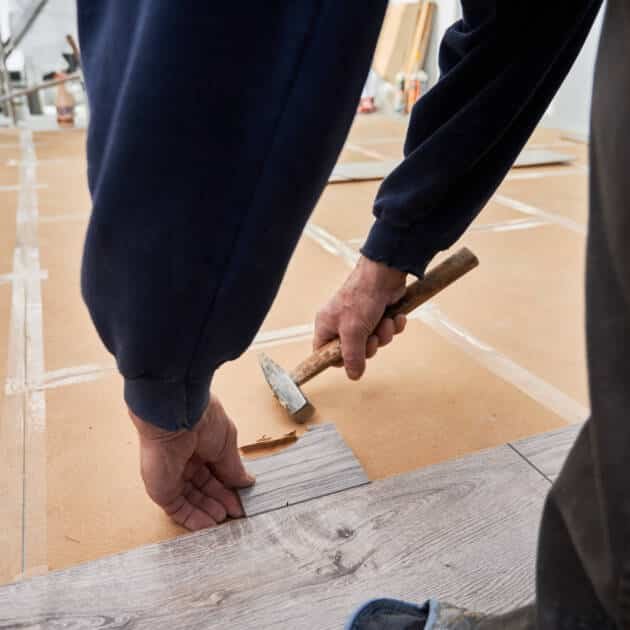 Close up of man construction worker using hammer tool while installing laminate flooring in apartment under renovation. Male hands laying laminate wooden panel on the floor. Home renovation concept.