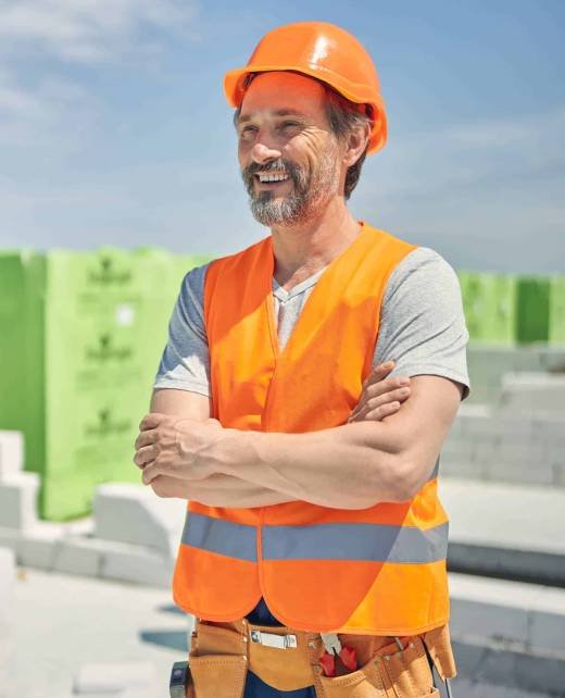 Merry mature bearded Caucasian builder standing outdoors Front view of a high-spirited grey-haired male worker with folded arms looking into the distance